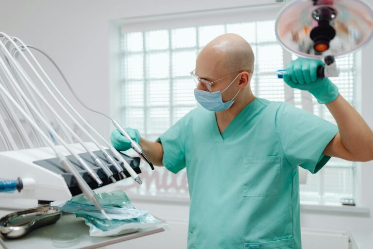 A bald dentist wearing green scrubs and a face mask adjusting dental equipment in a clinic setting.